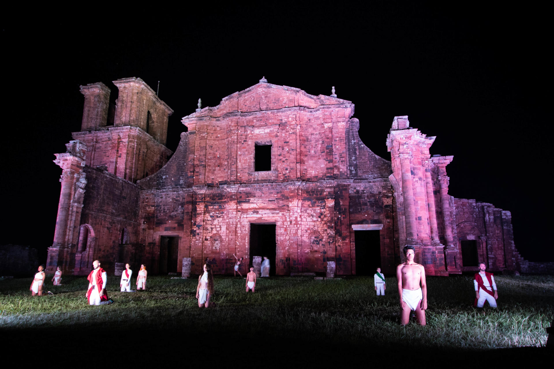 Foto colorida horizontal mostra cena do espetáculo, com os dançarinos em ação em frente às ruínas iluminadas de rosa à noite