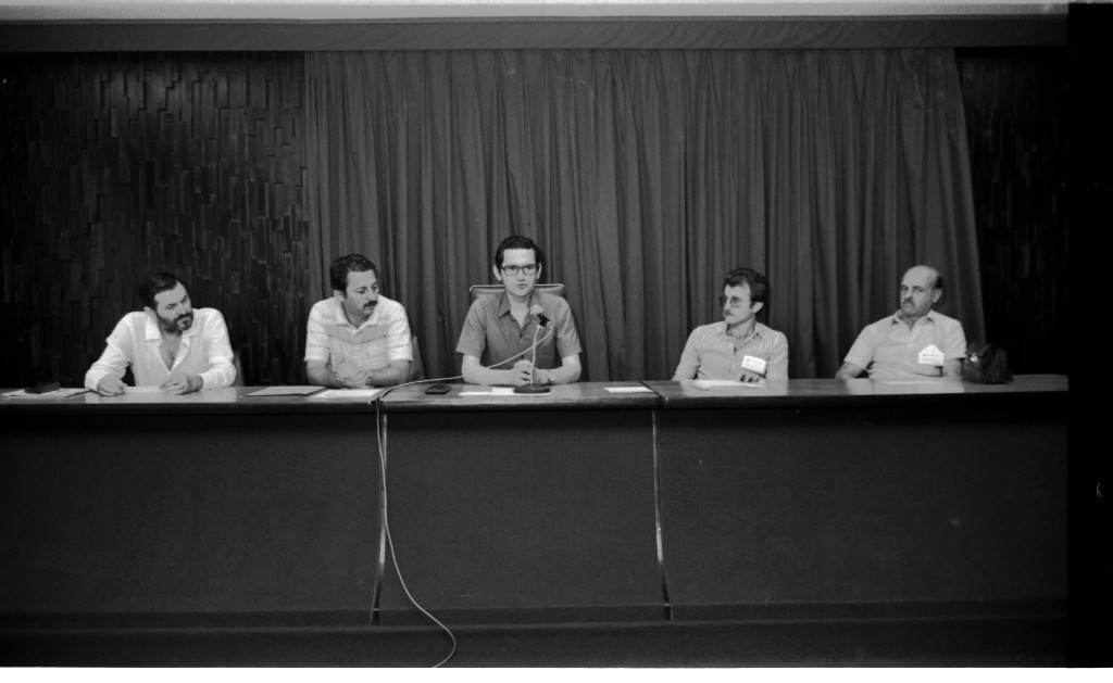 Foto horizontal preto e branca de palco com bancada e 5 homens sentados atrás da bancada.