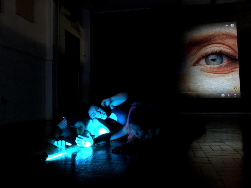 Três atores, um homem e duas mulheres, estão no centro de uma sala escura, deitados de bruços e iluminados por uma pequena luz azul à sua frente. Atrás dos atores uma projeção da imagem de um olho azul humano.