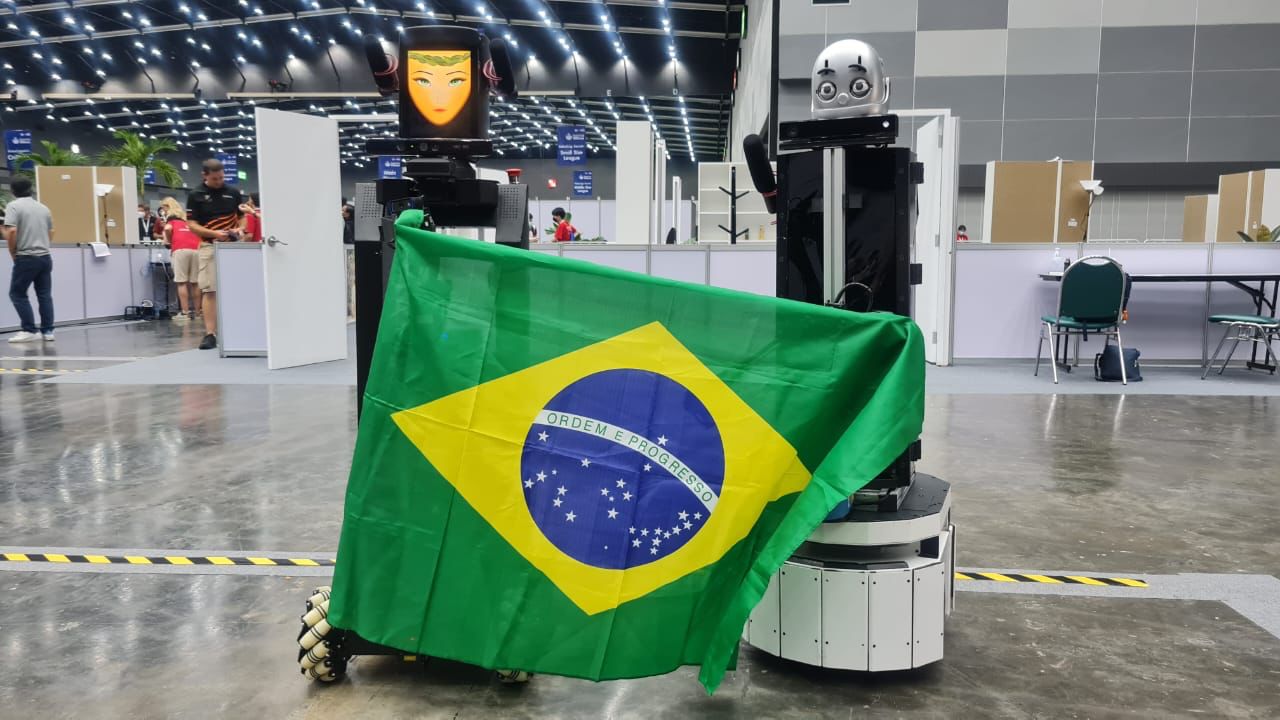 Foto, em formato horizontal e colorida. Robô da equipe RoboFEI, da FEI-SP e robô DoRIS, da equipe gaúcha FBOTS, respectivamente, segurando a bandeira do Brasil.