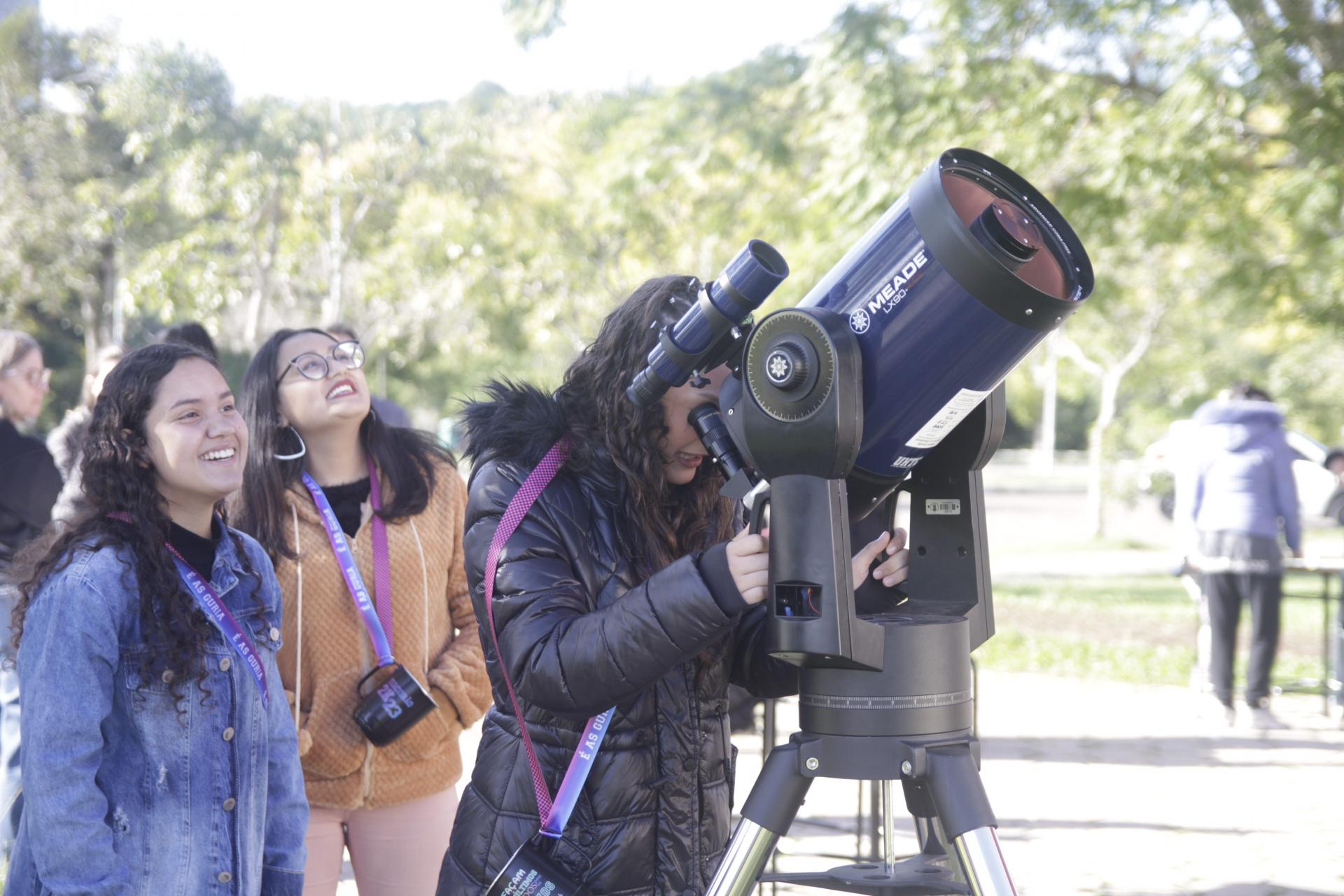 foto colorida horizontal com uma moça olhando por um telescópio, enquanto outras a observam e olham para cima, ao ar livre em um dia de sol