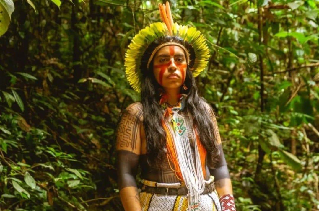 foto colorida horizontal com uma indígena, de cocar, adereços e pintura indígena, ao fundo uma floresta