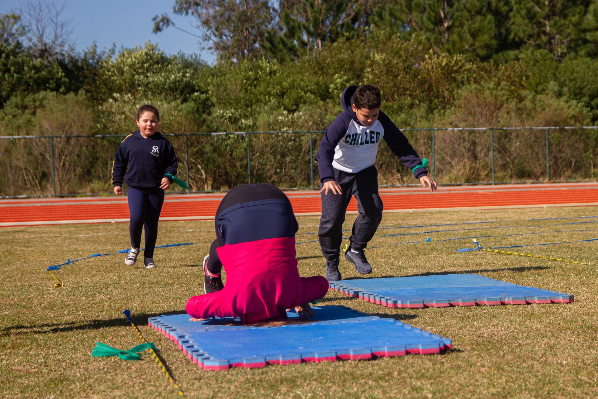 foto colorida horizontal de 3 crianças em um campo gramado, elas estão praticando atividades físicas, uma delas dá uma cambalhota sobre um tapume azul