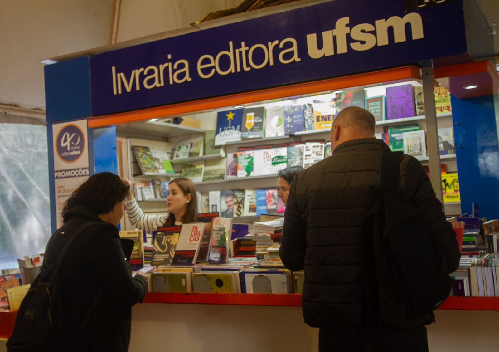 foto colorida horizontal com duas pessoas de costas em frente a uma banca da editora e livraria UFSM na feira do livro