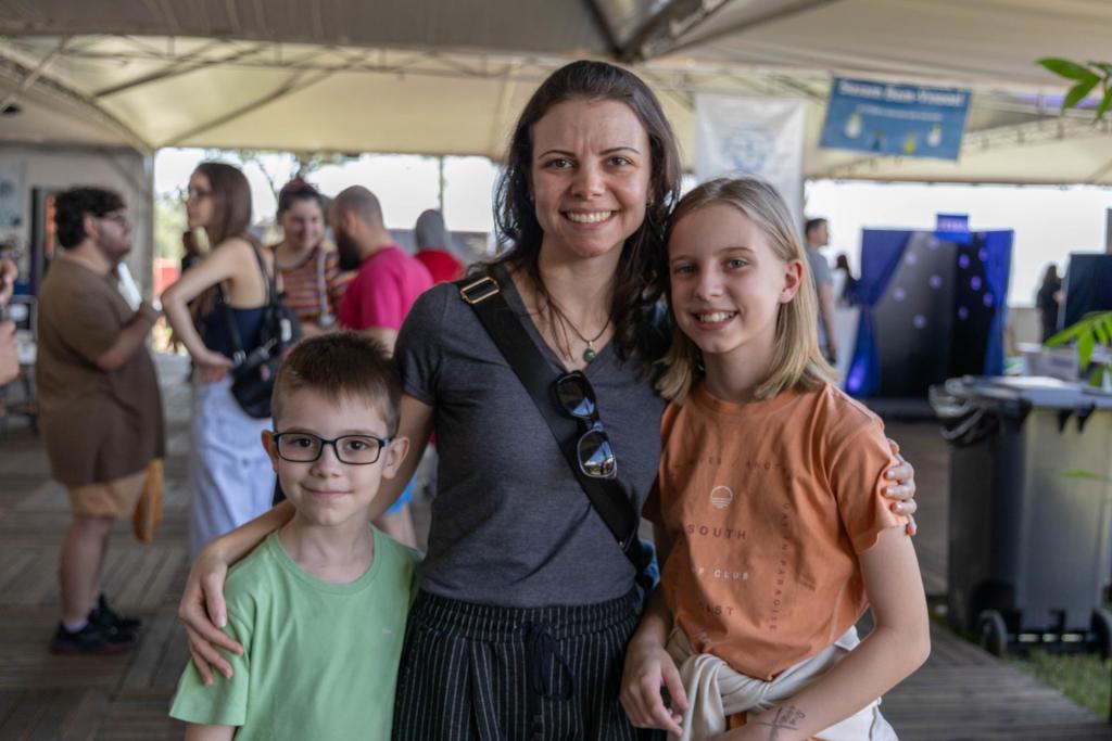 Foto colorida horizontal de mãe com dois filhos, um menino à esquerda e uma menina à direita. A foto mostra todos da cintura para cima