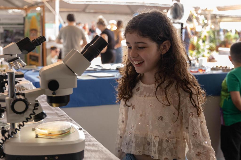 Foto colorida horizontal de menina sorrindo enquanto observa um microscópio