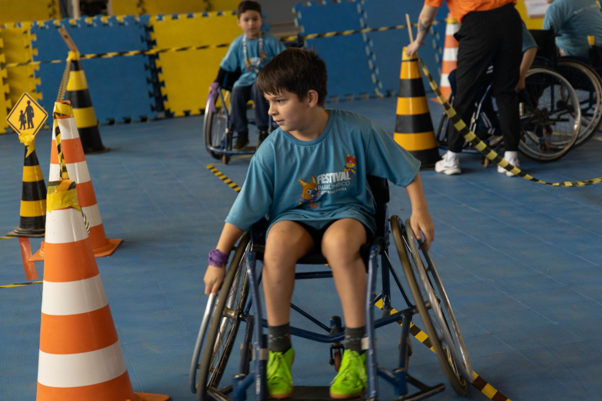 foto colorida em formato quadrado mostra um menino em uma cadeira de rodas participando de uma competição, em um espaço com cones