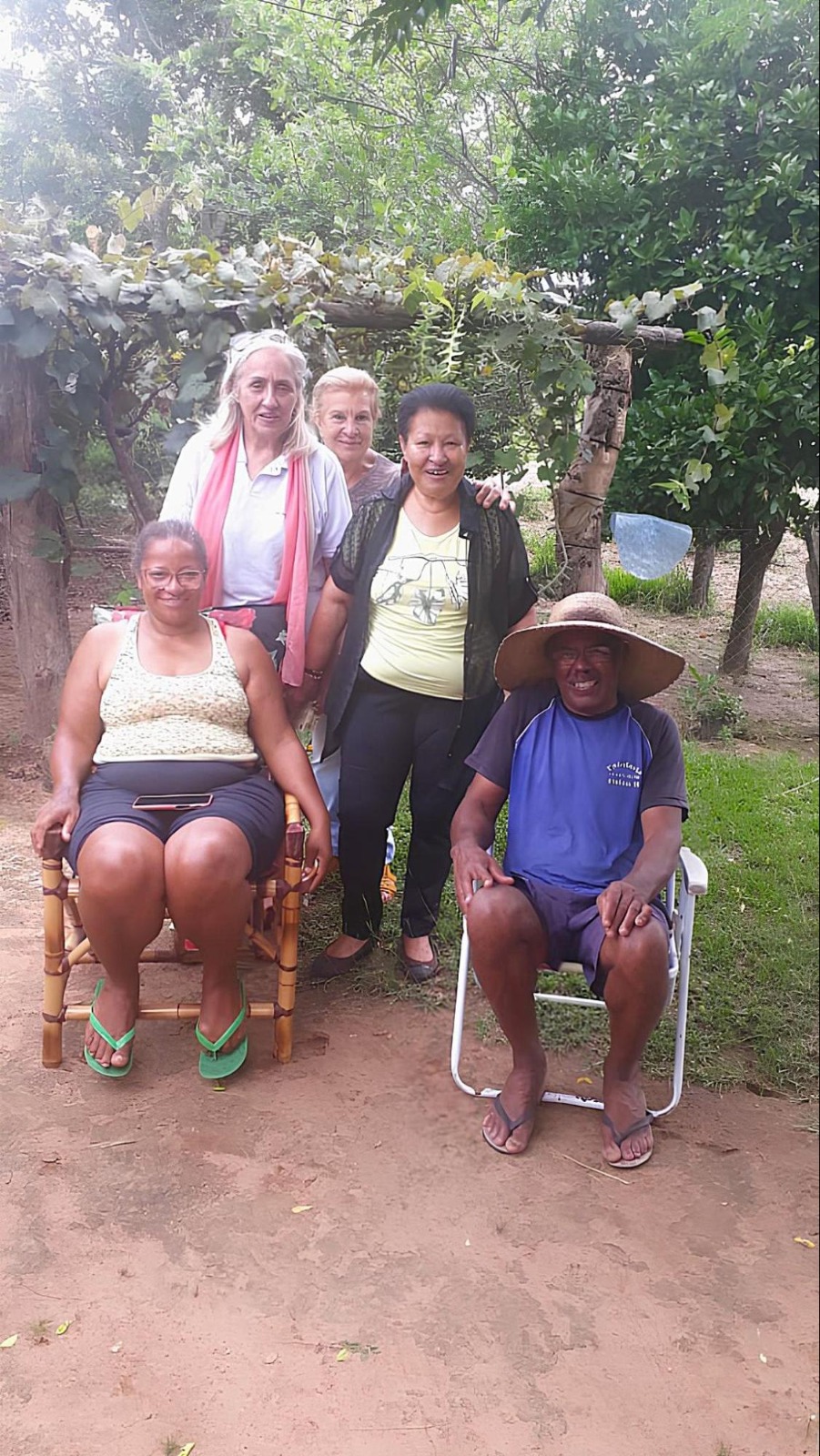 Foto colorida vertical em área externa rural, com chão batido e árvores atrás. A foto mostra cinco quatro pessoas, duas sentadas e três em pé. Todos posam para a foto.