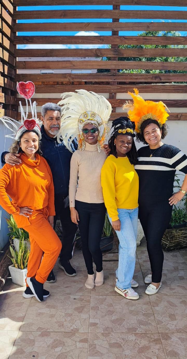 Foto colorida vertical de cinco pessoas em pé, sendo quatro mulheres e um homem. As mulheres usam adereços de carnaval na cabeça, como cocares com plumas. 