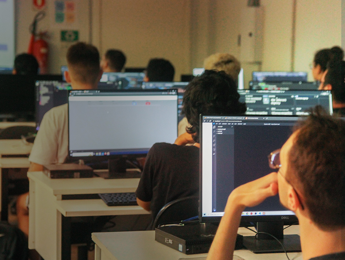 foto colorida quadrada de pessoas sentadas em frente a computadores em um ambiente que parece uma sala de aula. As pessoas aparecem de costas e de lado