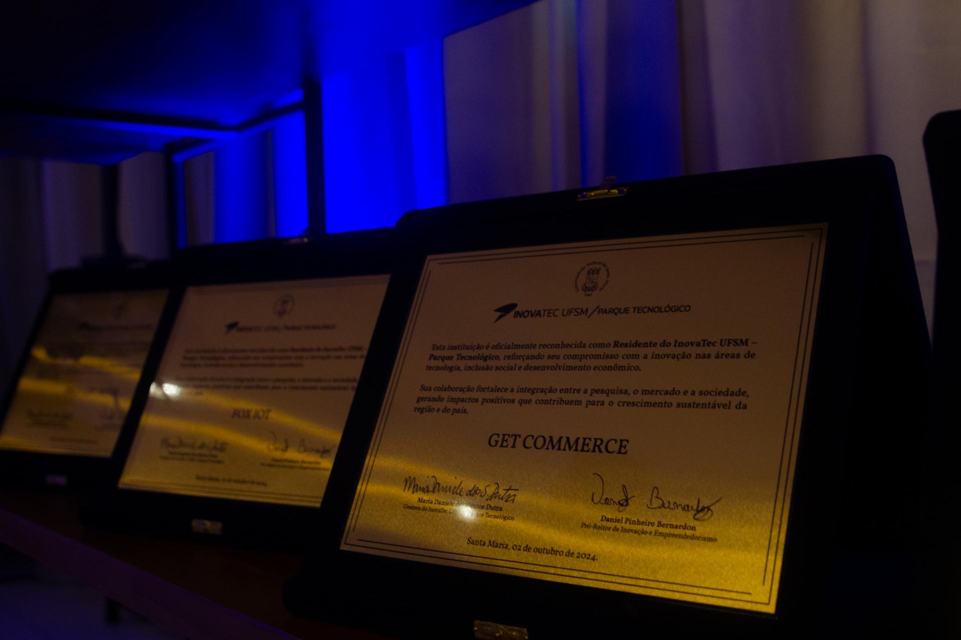 foto colorida quadrada mostra em destaque placas de homenagem, elas estão sob uma luz amarelada em um ambiente escuro, ao fundo uma luz azul