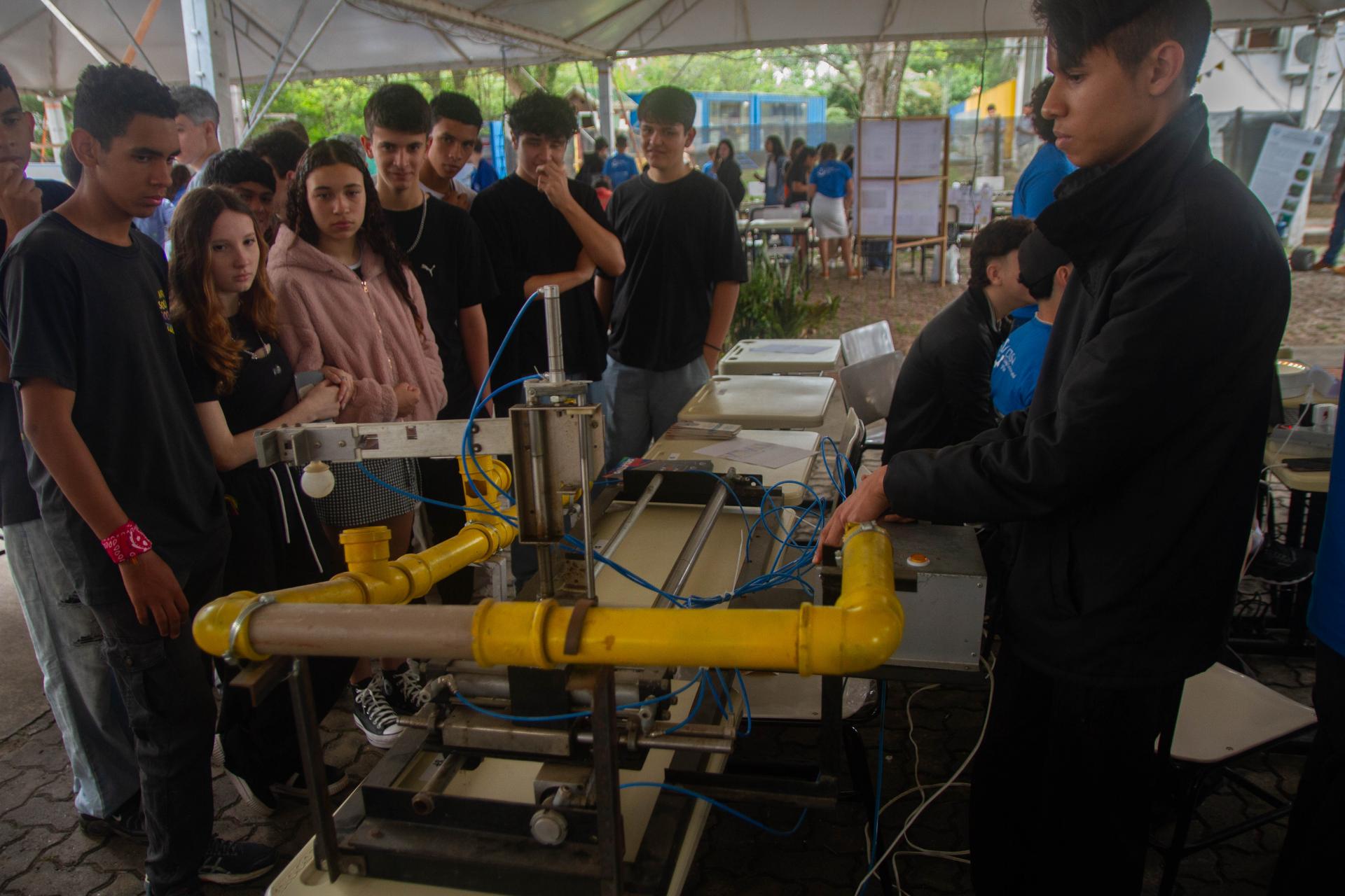 Foto colorida horizontal de feira. Do lado esquerdo, vários estudantes observando um equipamento. Do lado direito, um estudante em pé mostra como funciona a máquina.