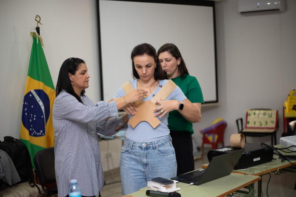 Foto colorida horizontal de três mulheres em pé em uma sala de aula. Uma delas usa coloca um colete e as outras duas a ajudam a ajustar. Atrás delas, uma bandeira do Brasil está hasteada