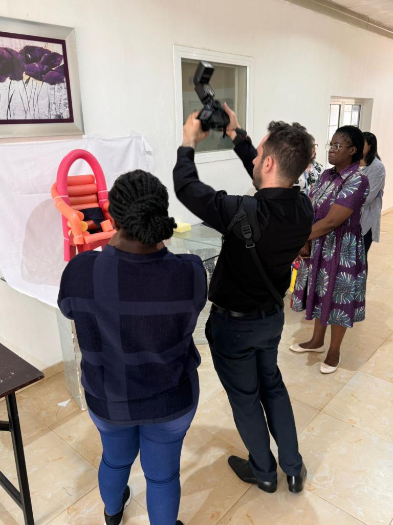 Foto colorida vertical de homem com câmera ao alto registrando objeto. Ao lado dele, mulheres observam a fotografia sendo feita