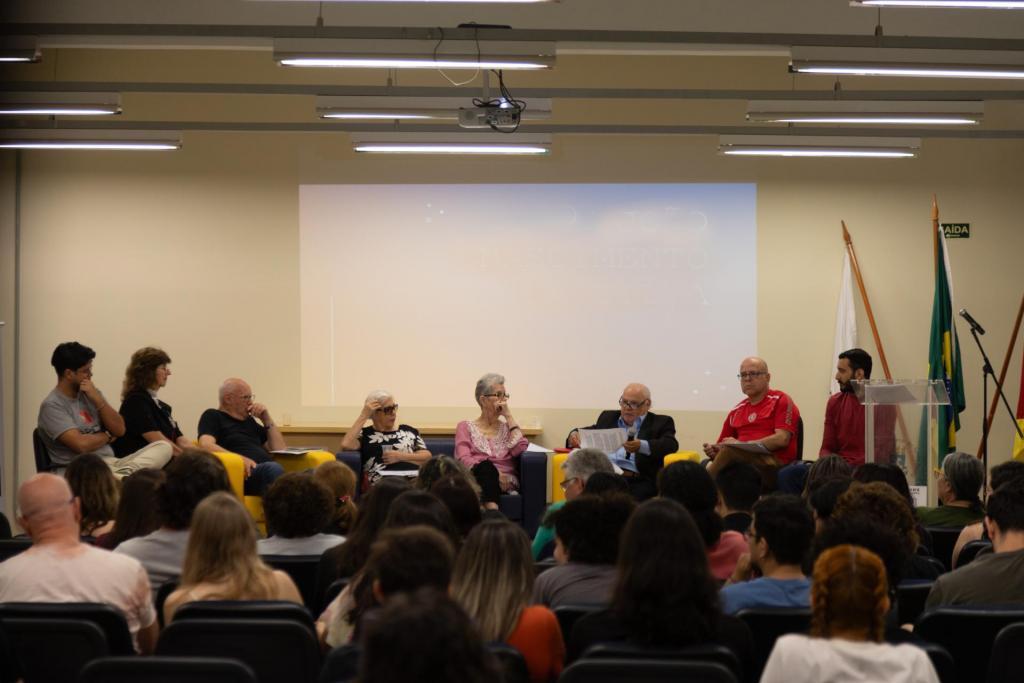 foto colorida horizontal de pessoas sentadas em semicírculo, no palco de um auditório, e de costas se vê pessoas assistindo à conversa