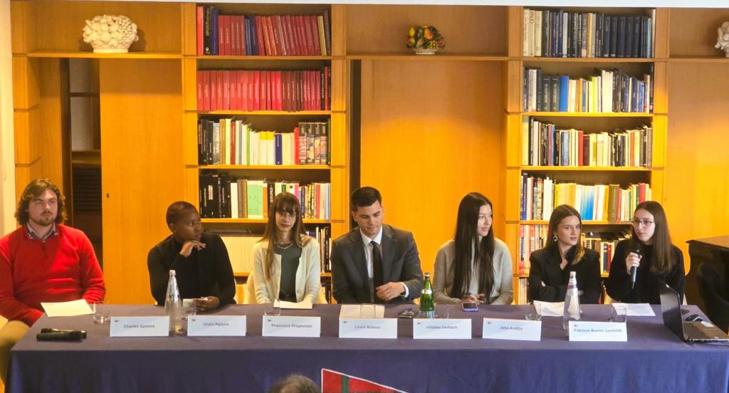 Foto colorida horizontal de sete jovens sentados em uma mesa de conferência. À frente de todos, placas com identificação dos nomes. Todos usam roupas formais. No canto à direita, estudante com microfone que representa a UFSM. Atrás de todos, uma parede amarela e duas estantes com livros até o teto