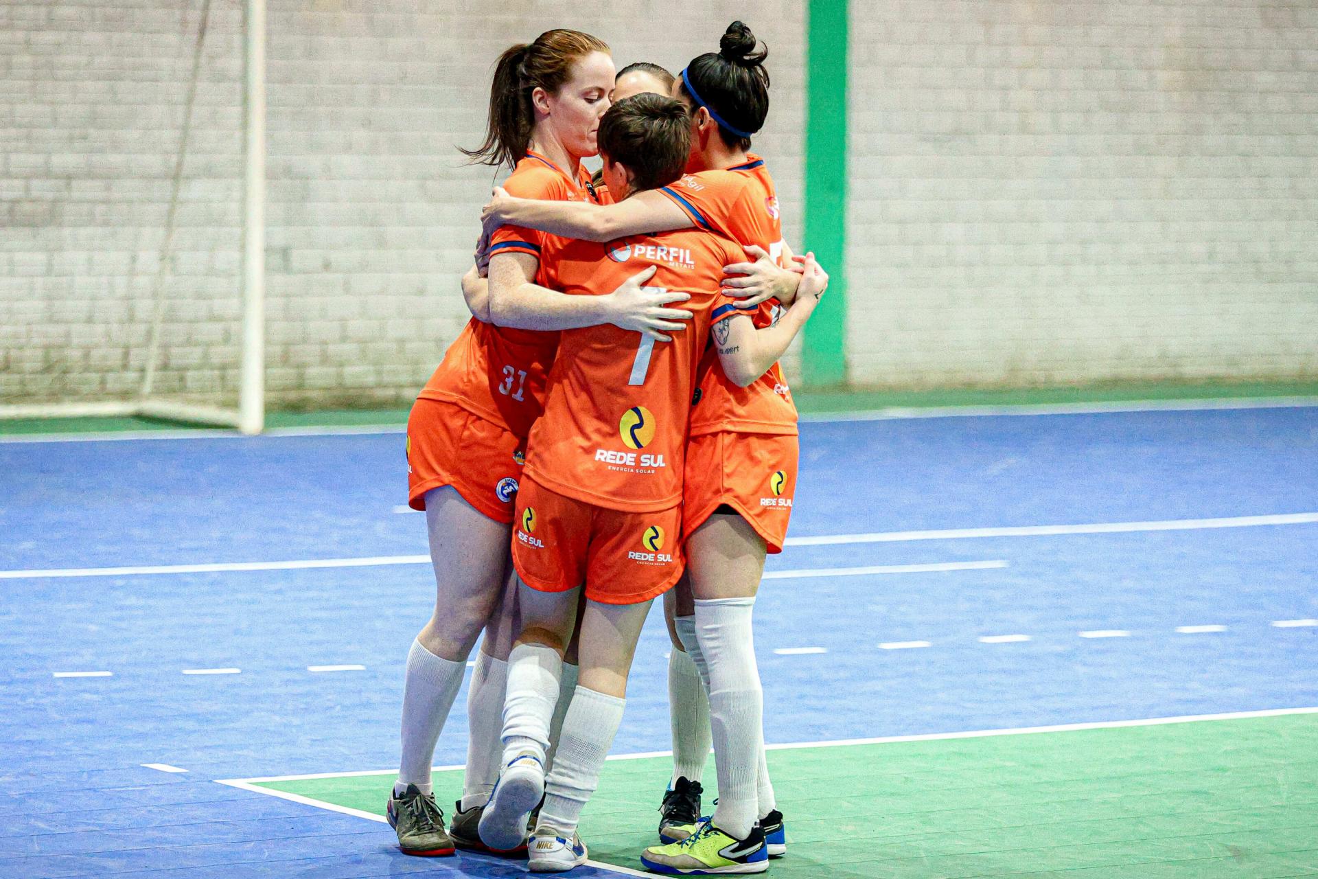 Foto colorida horizontal de quatro jogadoras, com uniformes laranjas, chuteiras e meias próximas ao joelho. Não é possível identificar o nome da equipe. As quatro estão abrançadas em círculo entre elas à frente de uma goleira de futebol em um ginásio fechado