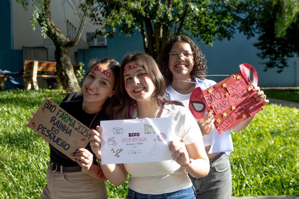 Foto colorida horizontal de três amigas segurando cartazes com mensagens de recepção. A foto mostra as três da cintura para cima em um gramado