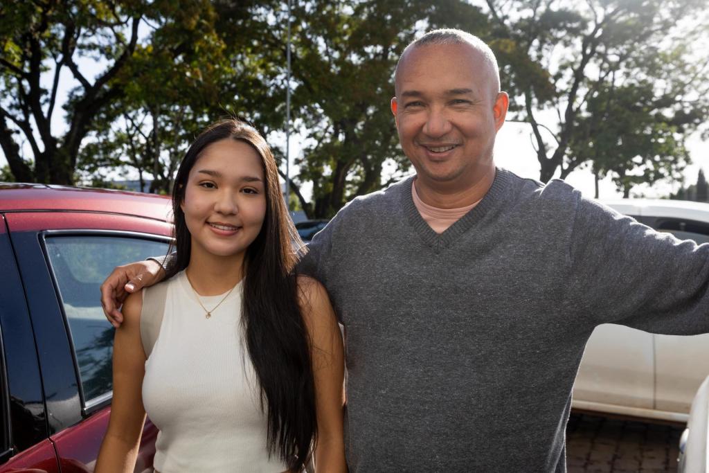 Foto colorida horizontal de jovem com seu pai. Os dois estão abraçados em frente a um estacionamento de carros.