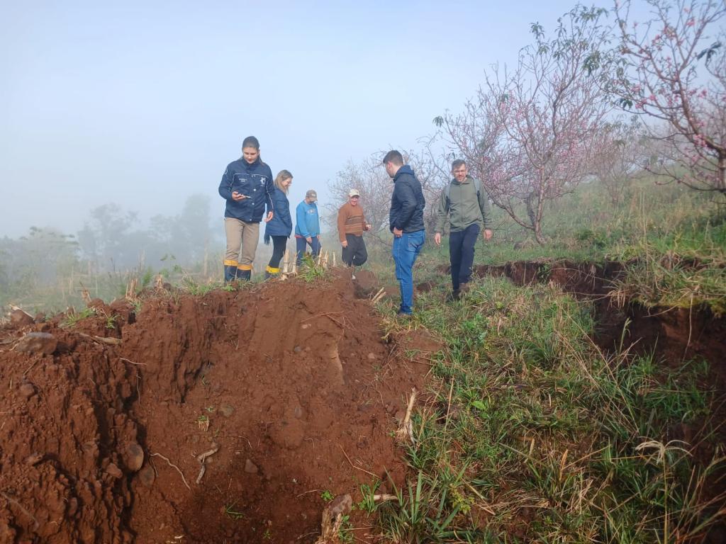 Foto colorida de equipes da UFSM em área rural de Agudo. Os membros da equipe analisam uma valeta no solo