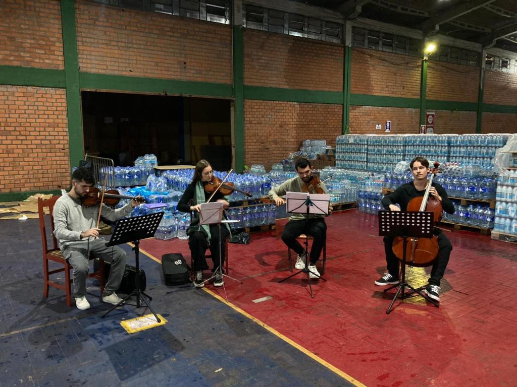 Foto colorida de grupo da orquestra se apresentando em abrigo de Santa Maria. Na imagem, quatro músicos aparecem tocando seus instrumentos com doações e mantimentos ao fundo