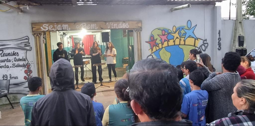 Foto colorida de pessoas assistindo a apresentação de músicos da orquestra em abrigo de Santa Maria