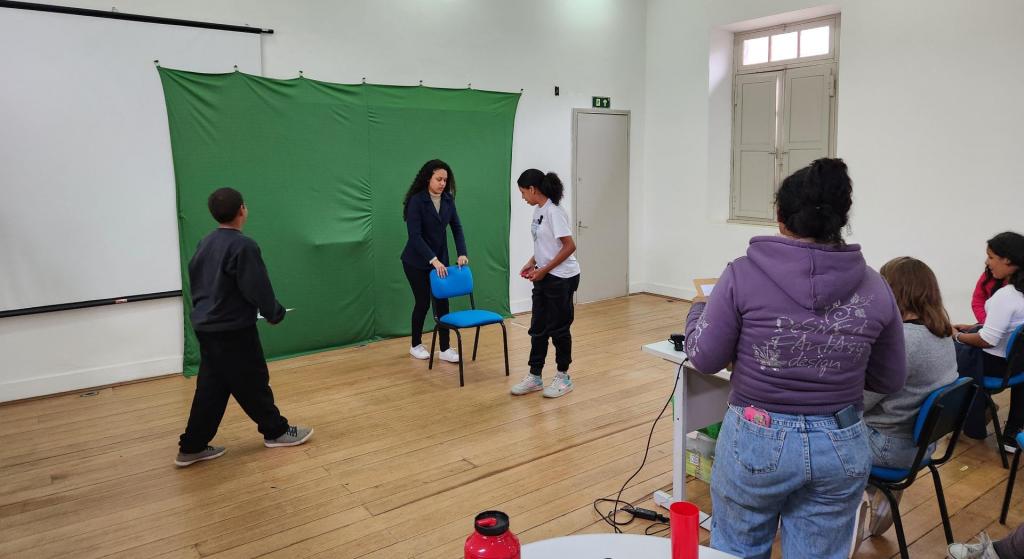 Foto de atividades de extensão com estudantes em sala de aula. No centro da imagem, professora segura cadeira para estudante. Ao fundo delas, aparece um chromakey verde