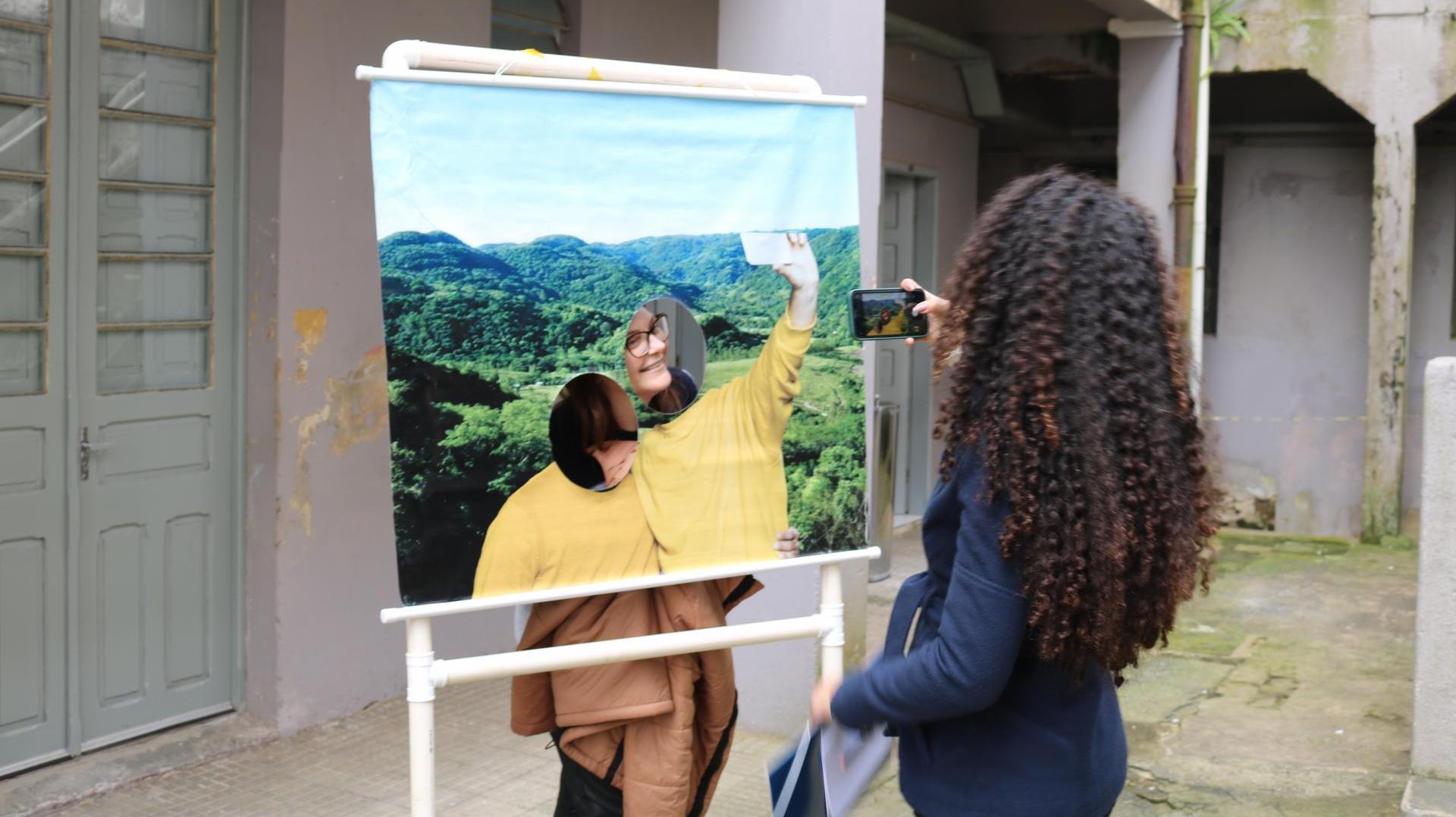 Foto colorida de estudantes participando de atividade de extensão. No centro da imagem, uma estudante aparece atrás de uma moldura do parque natural de Santa Maria. Ela sorri e olha para outra estudante que segura uma câmera