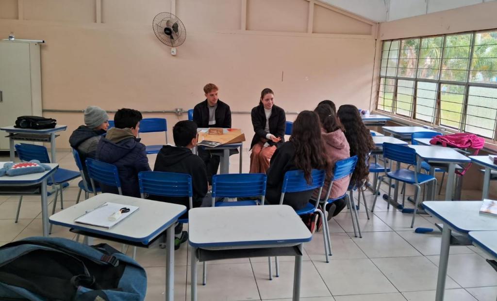 Foto colorida de estudantes sentados em uma semicírculo em uma sala de aula e conversando com dois membros do projeto