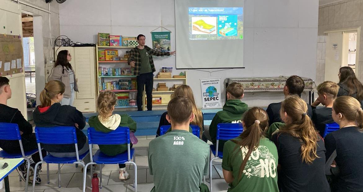 Foto colorida de atividade de extensão em escola de Paraíso do Sul. Na imagem, voluntário da UFSM aparece mostrando imagens em datashow para estudantes que estão de costas e sentados em uma sala de aula