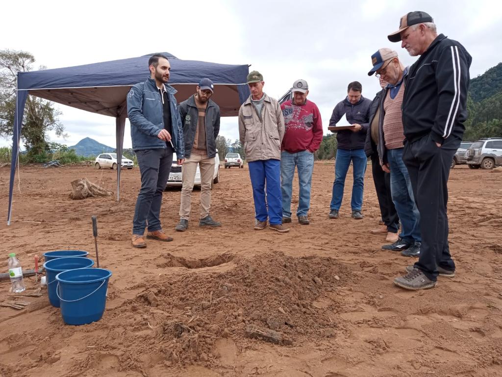 Foto colorida de Dia de Campo em Agudo. Na imagem, professor da UFSM explica detalhes sobre técnica de recuperação dos solos para agricultores locais. Eles estão de pé e diante de um montante de terra remexida com mudar de árvores ao lado.