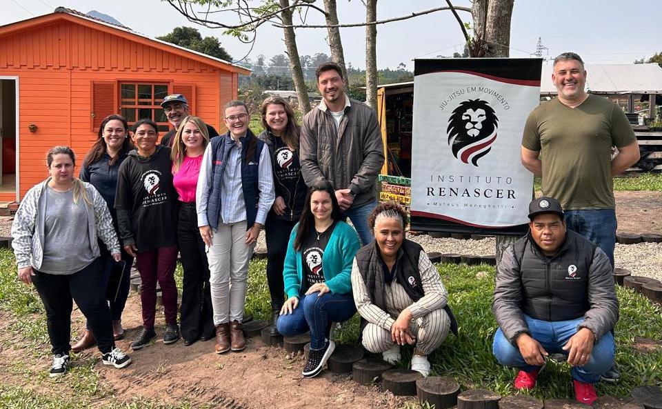 Foto colorida de reunião entre equipe da UFSM e Instituto Renascer. Na imagem, aparecem 11 pessoas posando para a foto. Três estão de cócoras e os demais de pé. Ao fundo casas e ao lado de um deles banner do Instituto Renascer