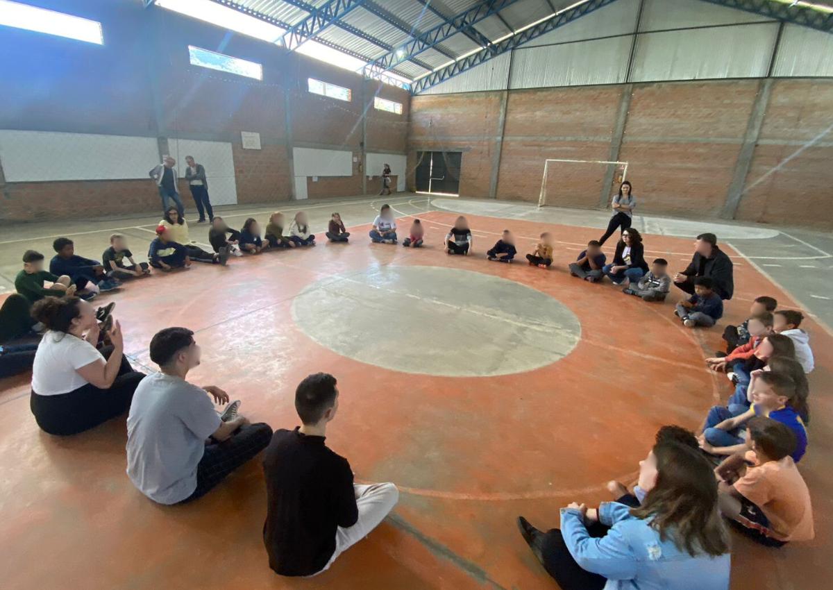 Foto colorida de estudantes de escola de Agudo sentados em circulo em ginásio de esportes