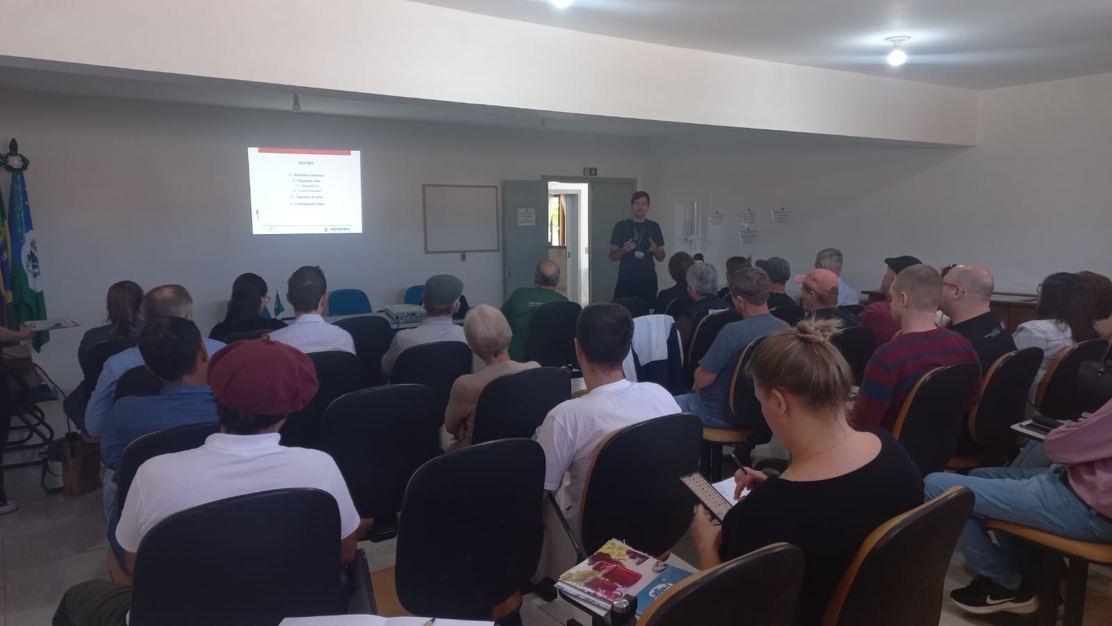 Foto colorida de palestra em Paraíso do Sul. Na imagem, diversas pessoas aparecem sentadas e olhando para uma parede com reprodução de data show. Professor da UFSM aparece ao fundo gesticulando.