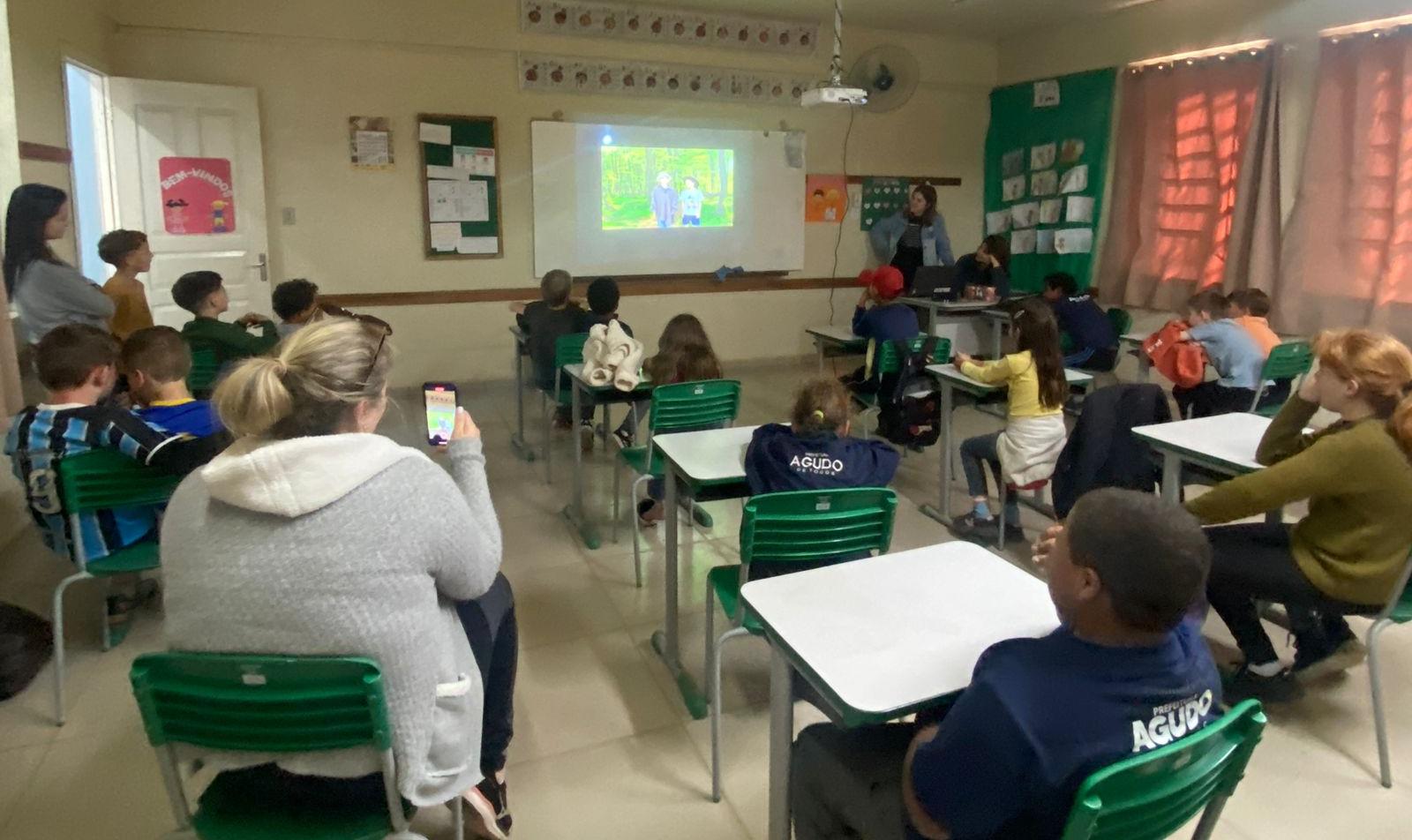 Foto colorida de ateliê em escola de Agudo. Na imagem, alunos aparecem sentados assistindo apresentação em data show no quadro da sala de aula.