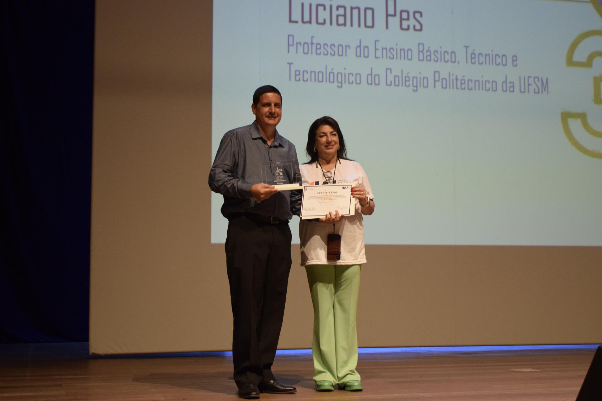 Foto colorida de Luciano Pes recebendo placa de reconhecimento da vice-reitora Martha Adaime