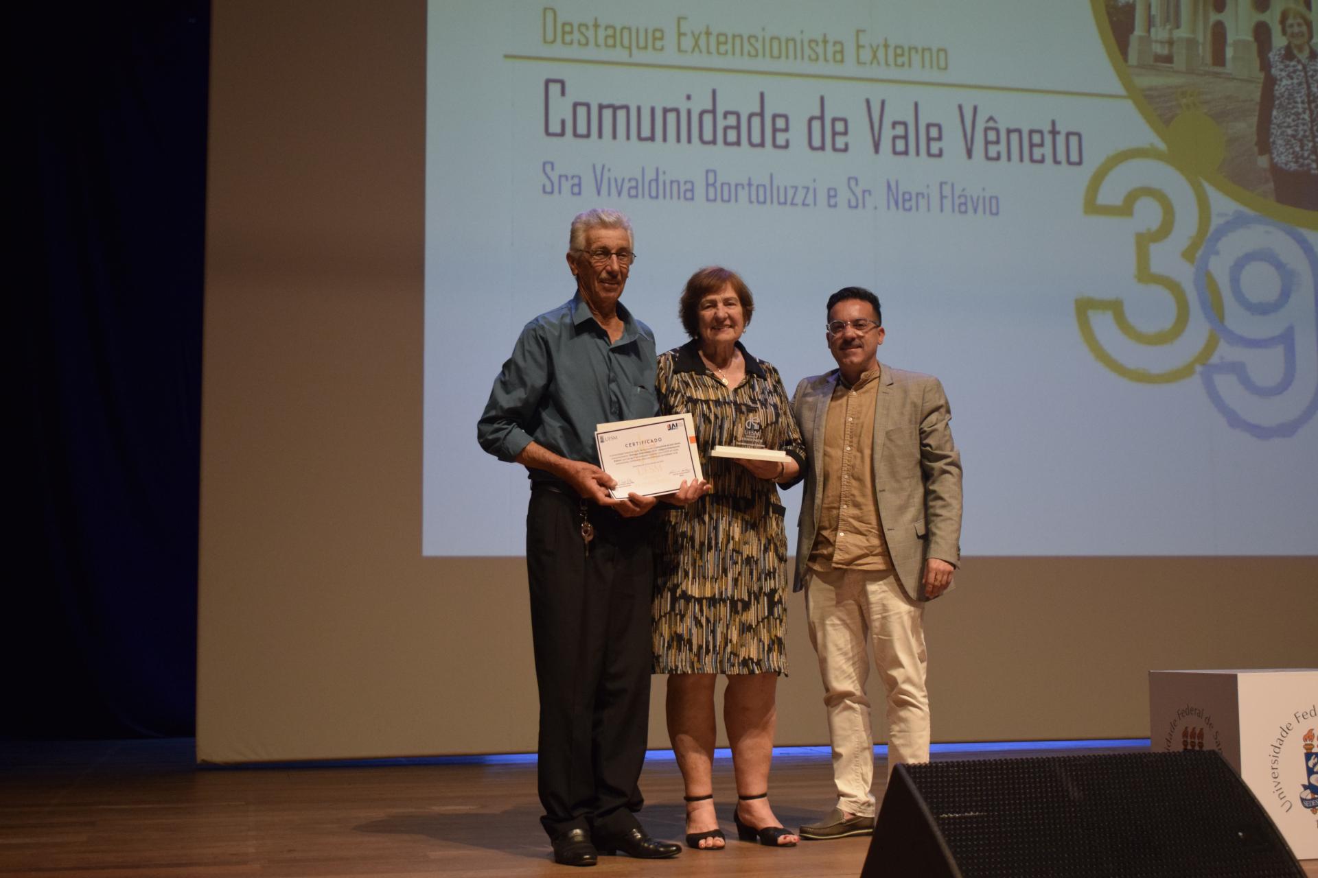 Foto colorida de Neri Flávio e, Vivaldina recebendo a placa de reconhecimento de Flavi Lisboa