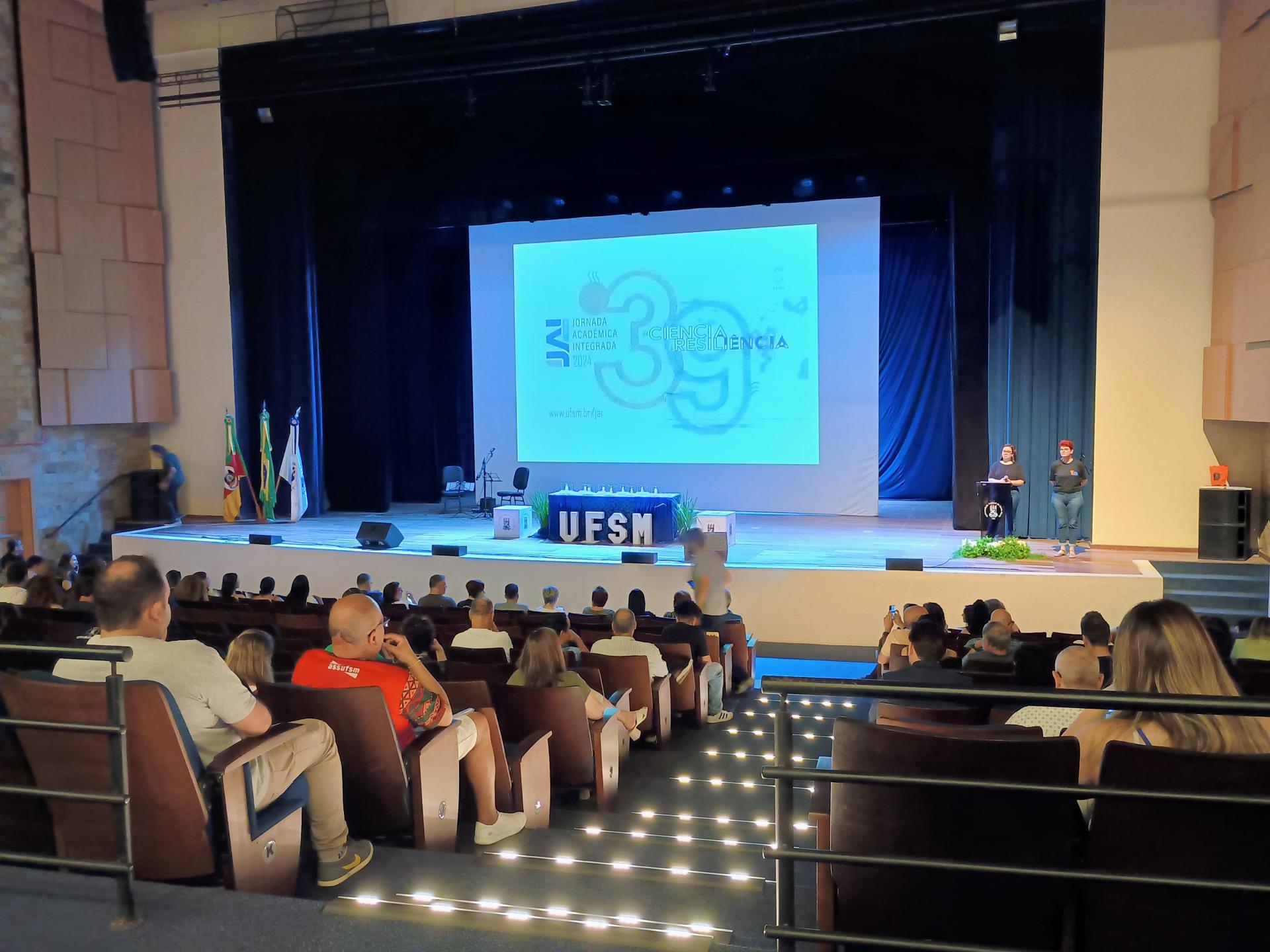 Foto colorida do Centro de Conveções da UFSM durante Abertura da JAI. Na imagem, aparece parte do público e o palco do evento.