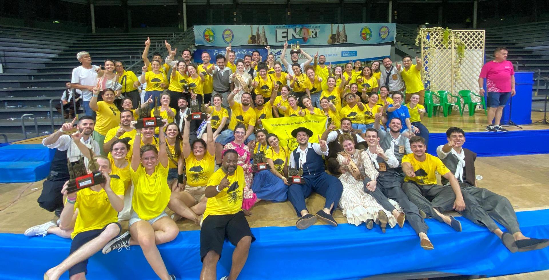 Foto colorida de grupo do DTG Noel Guarany comemorando premiação no Enart. Eles estão em um ginásio, a maioria com uma camisa amarela com símbolo do DTG e sorriem