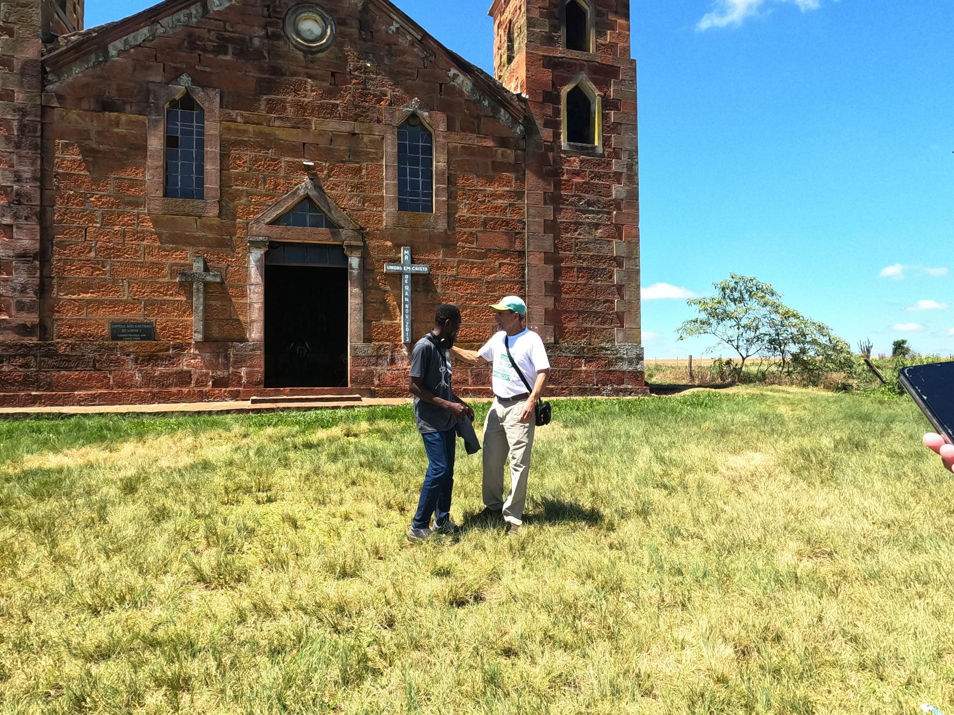 Foto colorida de duas pessoas conversando em frente da Igreja de Pedra São Caetano em Nova Esperança do Sul