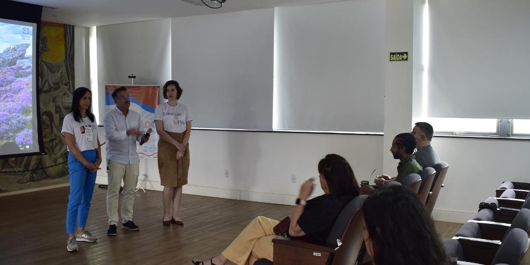 Foto colorida do Fórum Permanente em Santa Maria. Na imagem aparece o Pró-reitor Flavi, a pró-reitora adjunta Jaciele e outra mulher em pé, falando e olhando para o público