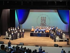 Foto retangular do palco do Centro de Convenções com os formandos, autoridades e homenageados em posição de respeito à execução do Hino Nacional Brasileiro 