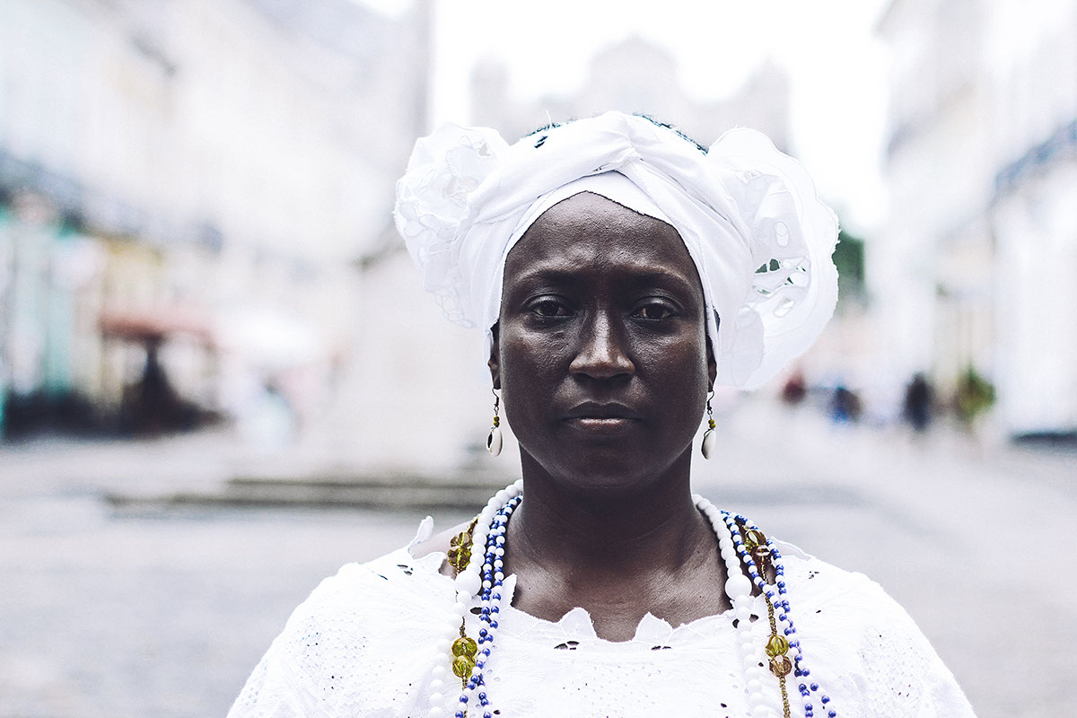 Uma mulher preta, com vestimentas brancas que remetem a cultura religiosa da umbanda. Ela está no centro da fotografia, e foi retratada dos ombros para cima, com enfoque no rosto. O fundo está desfocado.