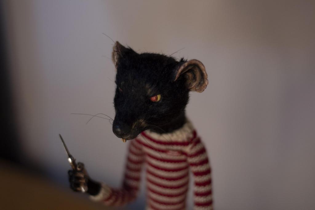 Fotografia horizontal e colorida de uma miniatura do personagem "Rat" do filme "O Fantástico Sr. Raposo". O rato está em pé sob uma mesa de madeira. O rato tem pelagem preta, olhos vermelhos e orelhas grandes arredondadas. O rato veste um blusão listrado de tricot nas cores branca e vermelha. Na mão esquerda a miniatura segura uma faca. O fundo é uma parede branca com sombras escuras.