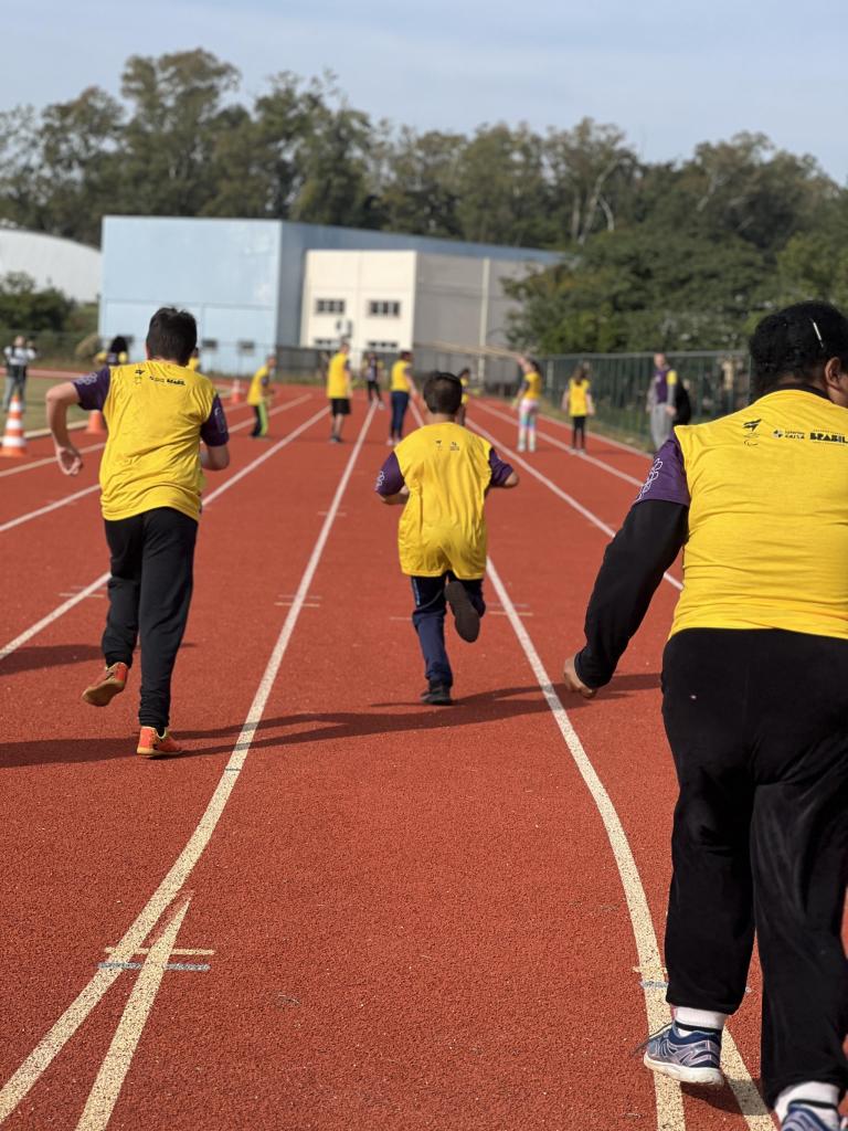 Foto na vertical. Em quadra de corrida na cor vermelha. Três crianças em destaque de costas com camiseta amarela com mangas roxas. Crianças correndo em direção a mais alunos que estão esperando ao fundo. Prédio azul ao fundo, em desfoque. 