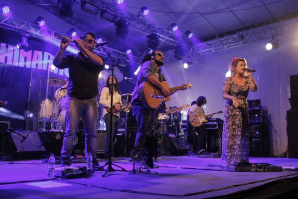 Fotografia horizontal, colorida e em plano geral de uma banda que se apresenta em um palco. O grupo musical é composto por seis integrantes, cinco homens e uma mulher, que é a vocalista. No palco, a formação da banda está dividida com três pessoas na parte da frente e as outras três na parte de trás.Na frente,um homem de estatura média está em pé,de óculos escuro, segurando uma flauta, com 2 garrafas de água ao lado dele, ao lado dele temos outro homem também de óculos escuro, porém com uma compleição física mais larga, cabelos longos, tocando um violão, a sua direita temos uma mulher que está segurando um microfone e está cantando num palco. Ela possui uma flor na cabeça e está com um vestido longo. Atrás temos um integrante com um violão menor, de cabelos longos e lisos de roupa clara, a sua direita um homem de cabelos longos, porém ondulados com uma guitarra bem grande. Ao fundo, há uma bandeira preta com o nome da banda Chimarruts escrito em branco.