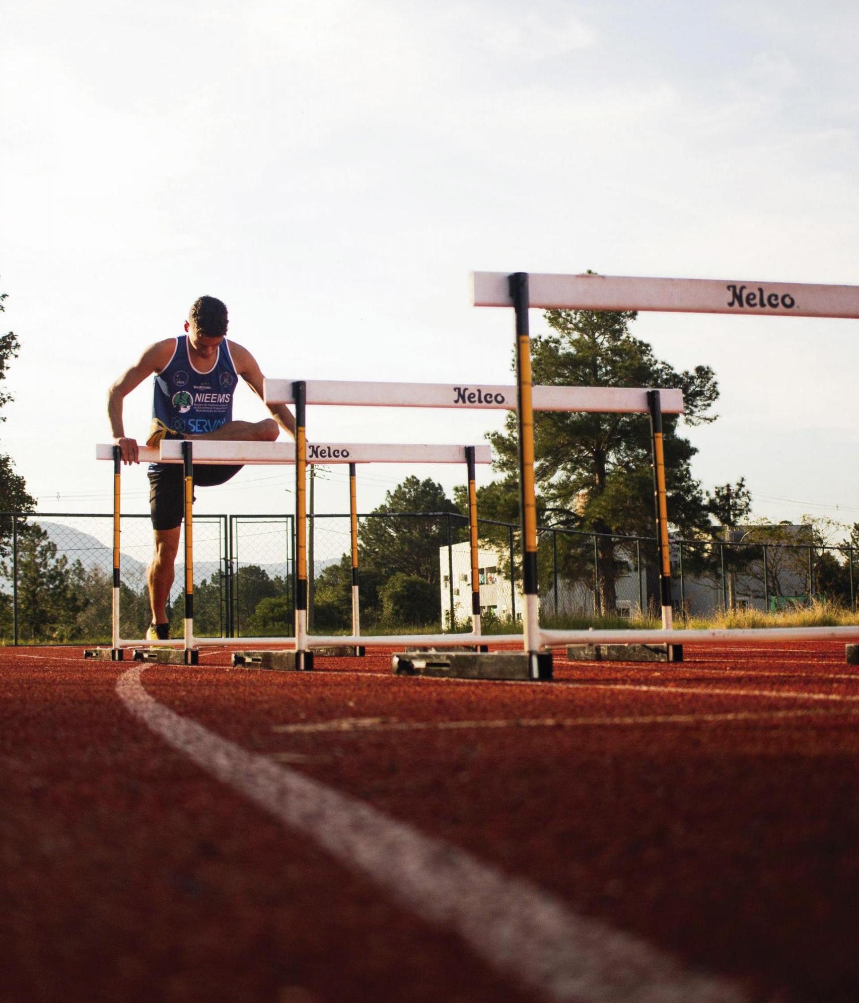 Fotografia vertical, colorida e em Contra-Plongée, de baixo para cima, com foco em um atleta que alonga a perna direita em uma barreira de atletismo na pista da UFSM. O homem tem pele branca, cabelos castanhos e curtos e olha para baixo. Ele usa uma regata azul de treino e um short preto. À sua frente estão mais três barreiras, que são formadas por duas barras verticais que sustentam uma barra horizontal. A pista é vermelha com listras brancas. O dia está ensolarado e ao fundo, há uma cerca, um prédio branco e árvores.