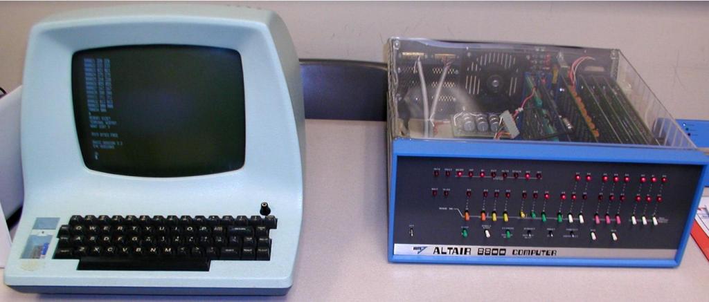 Imagem do kit do computador Altair 8800 com a parte superior em vidro mostrando seus componentes internos e seu painel frontal com uma série de leds e de botões interruptores. Acompanhando também o seu terminal composto pela união de uma tela com um teclado convencional embutido.