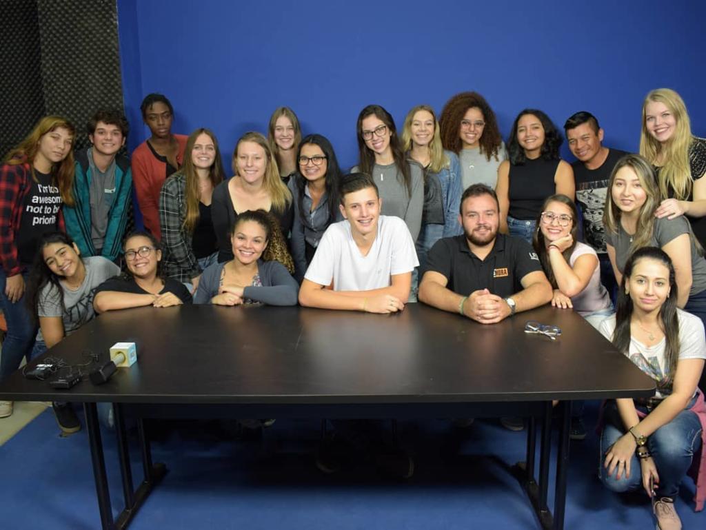 Foto horizontal com diversas pessoas atrás de uma mesa preta com fundo azul