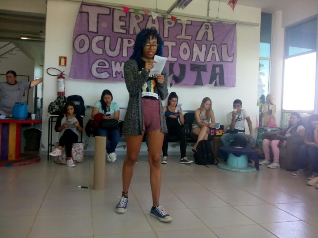 Mulher em pé falando no microfone, ao fundo pessoas sentadas e faiza lilás escrito Terapia Ocupacional em luta